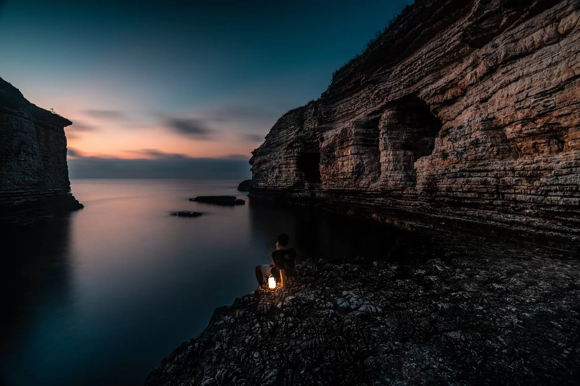 person sitting on sea coast among rocks at dusk